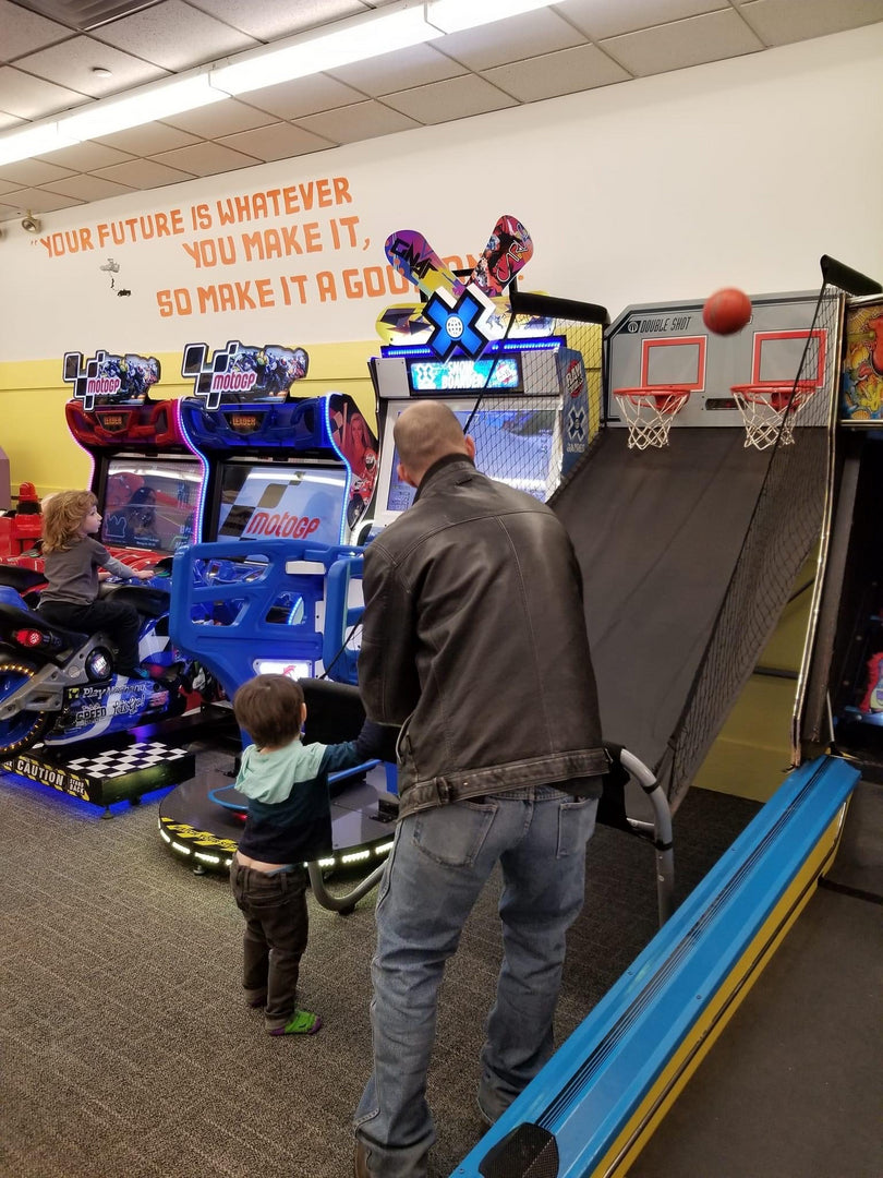 An adult playing a basketball game while two children play racing arcade games.